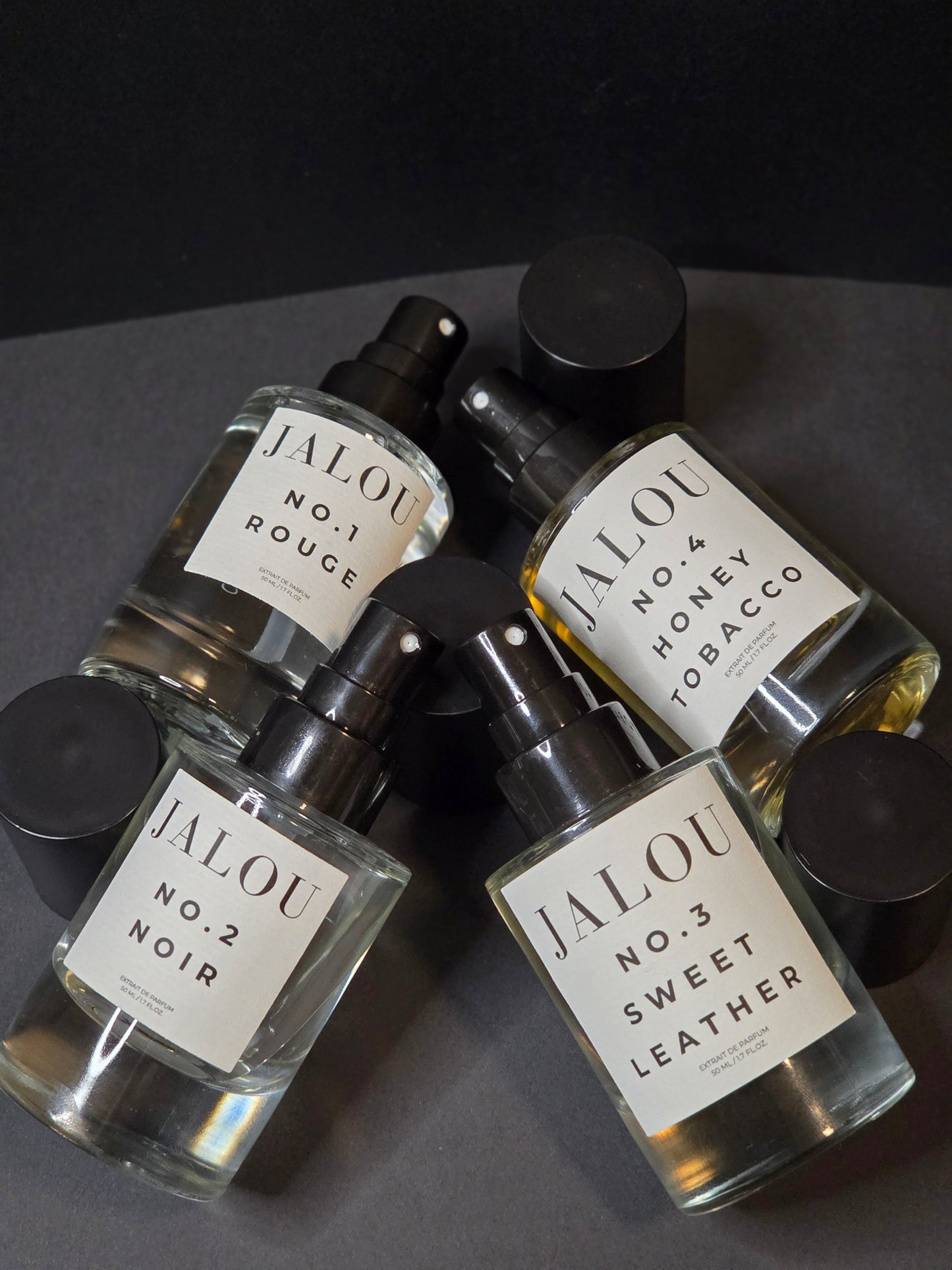 Vier Parfumflakons mit Sprühaufsatz, beschriftet mit No. 1 Rouge, No. 2 Noir, No. 3 Sweet Leather und No. 4 Honey Tobacco, auf dunklem Hintergrund.