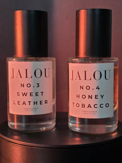Zwei JALOU Extrait de Parfum Flakons – NO. 3 SWEET LEATHER und NO. 4 HONEY TOBACCO – nebeneinander auf einem dunklen Podest bei warmer, rötlicher Beleuchtung