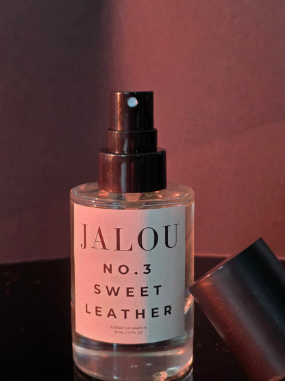 Nahaufnahme eines JALOU NO. 3 SWEET LEATHER Flakons bei warmer, rötlicher Beleuchtung mit nebenliegendem Deckel vor einem dunklen Hintergrund.