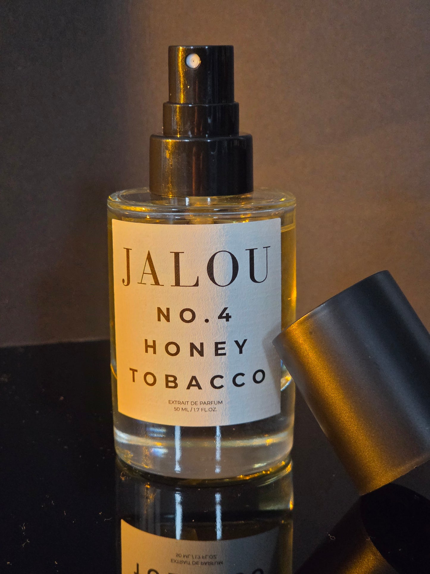 Ein Flakon JALOU NO. 4 HONEY TOBACCO auf einer spiegelnden schwarzen Fläche mit danebenliegendem Deckel unter warmem Spotlight vor dunklem Hintergrund.