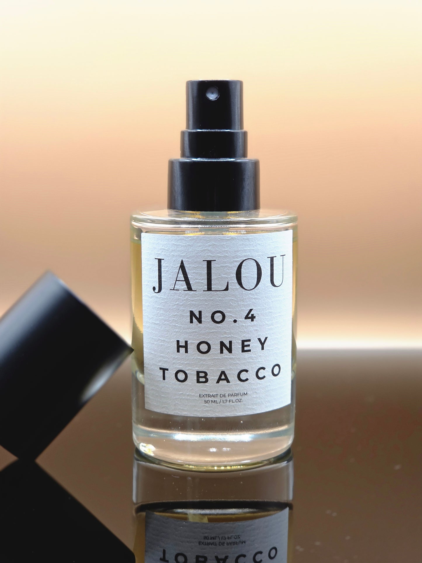 Ein Flakon JALOU NO. 4 HONEY TOBACCO mit abgenommenem Deckel auf einer spiegelnden Oberfläche vor einem warmen, hellen Hintergrund.