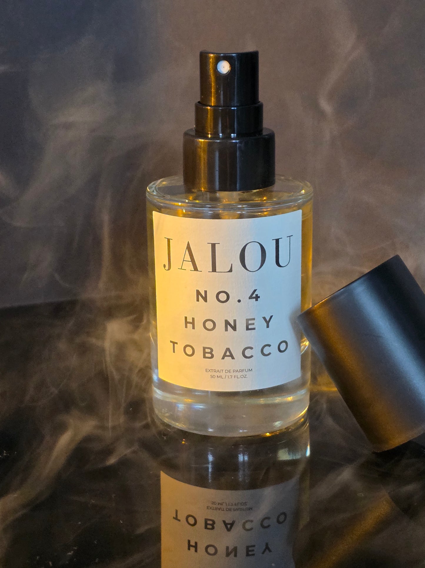 Ein Flakon JALOU NO. 4 HONEY TOBACCO auf einer spiegelnden Oberfläche, umhüllt von mystischen Rauchschwaden vor einem dunklen Hintergrund.